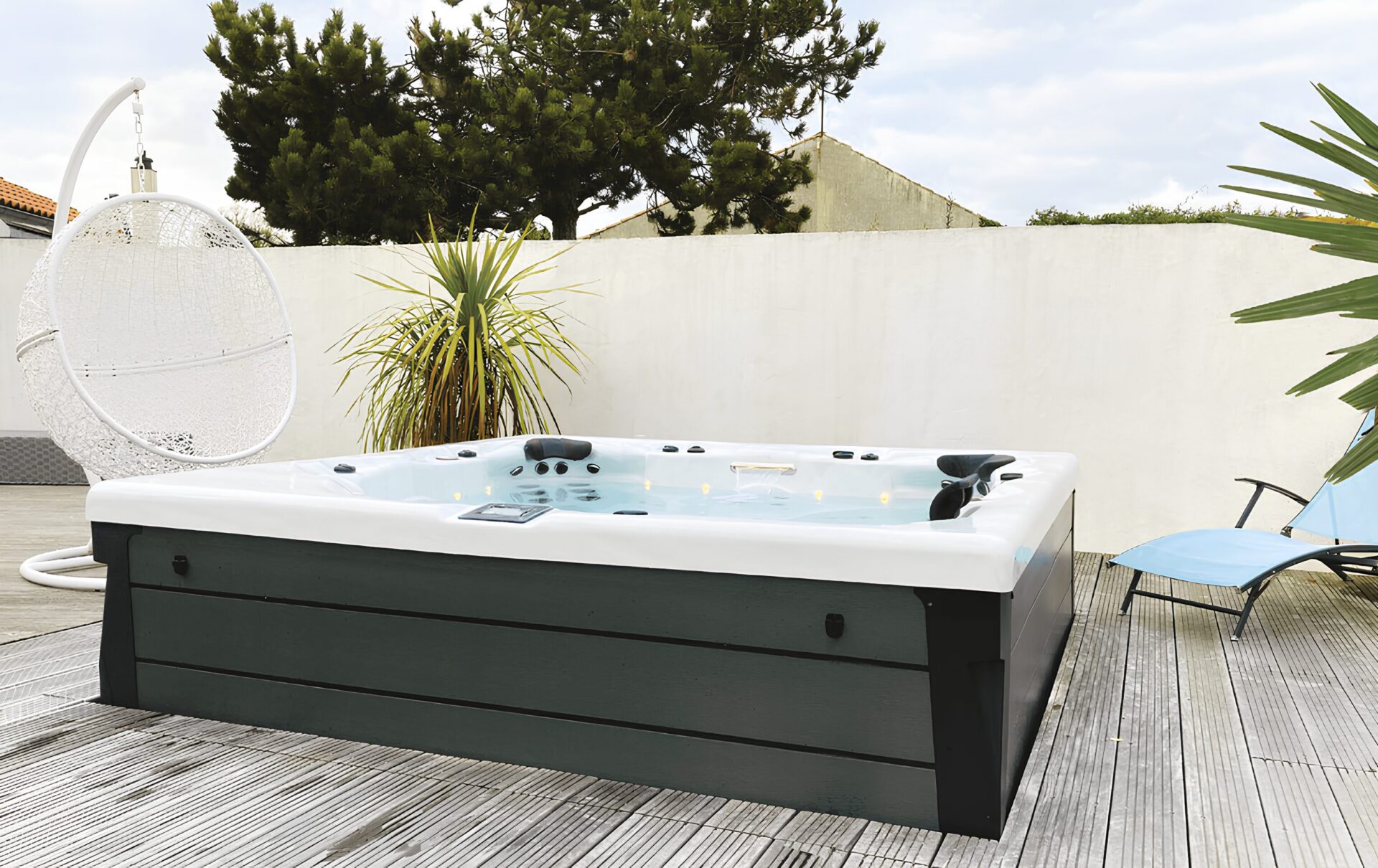 Jacuzzi extérieur sur terrasse en bois avec chaise longue bleue, chaise suspendue blanche et plantes.
