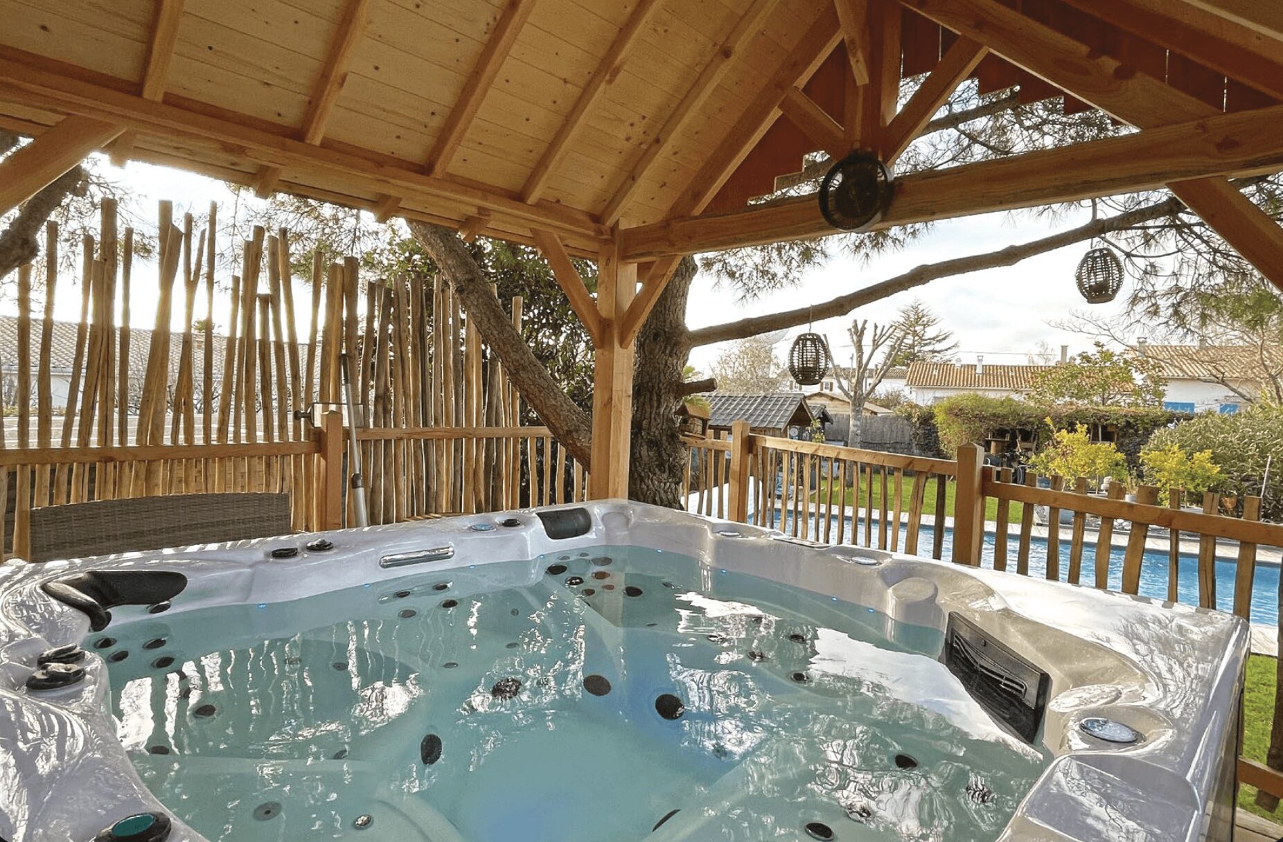 Jacuzzi sous pergola en bois avec piscine, arbres, buissons et maisons en arrière-plan.