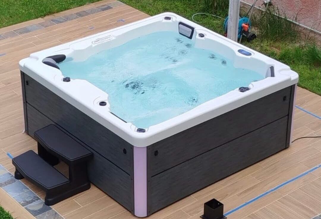 Spa carré extérieur en fonctionnement avec marches noires, sol carrelé imitation bois et pelouse.